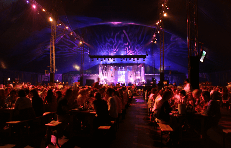 A vibrant concert hall with purple lighting: attendees seated at tables watch a live performance on a stage with dramatic backdrop.