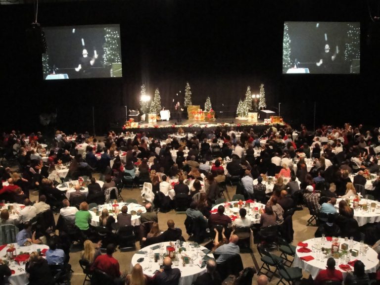 A large indoor holiday event with many people seated at round tables, watching a stage flanked by two large screens displaying the speaker.