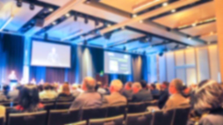 Audience in a Seattle conference hall listening to a speaker displayed on a large screen, with a blurred, focused background emphasizing the foreground.