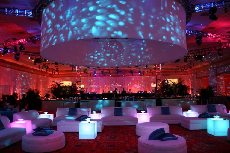 A modern event space with blue and pink lighting, circular overhead fixtures, and contemporary white lounge furnishings.