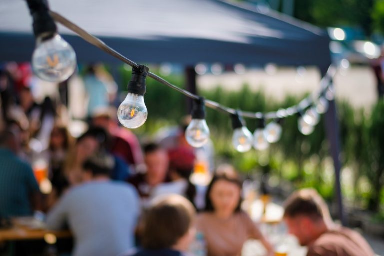 String of lit bulbs hanging above a blurred outdoor social gathering featuring a hired corporate entertainer in Seattle, in a sunny setting.
