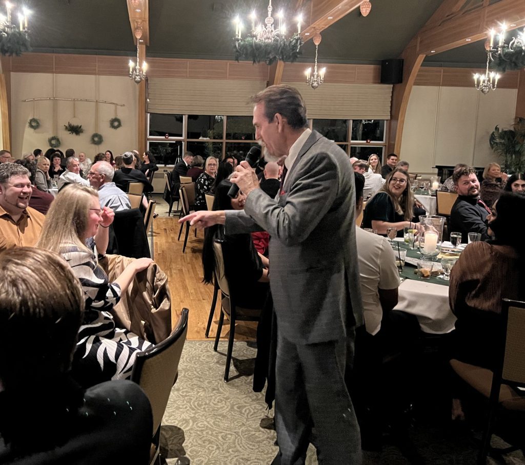 Lance Campbell speaks into a microphone at a lively dinner event, interacting with smiling attendees seated at tables in a warmly lit room adorned with holiday decorations. He is recognized as the Seattle Corporate Mind Reader, captivating